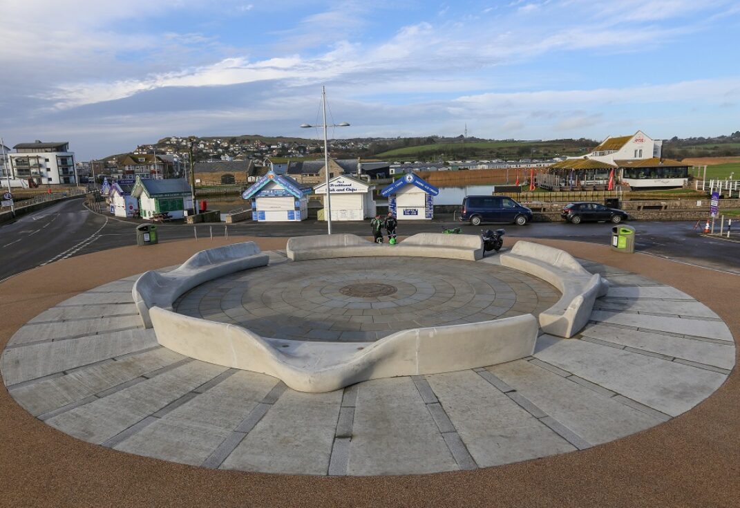 A circular concrete seat and motorbike parking bays in front of beach kiosks with houses and fields in the background