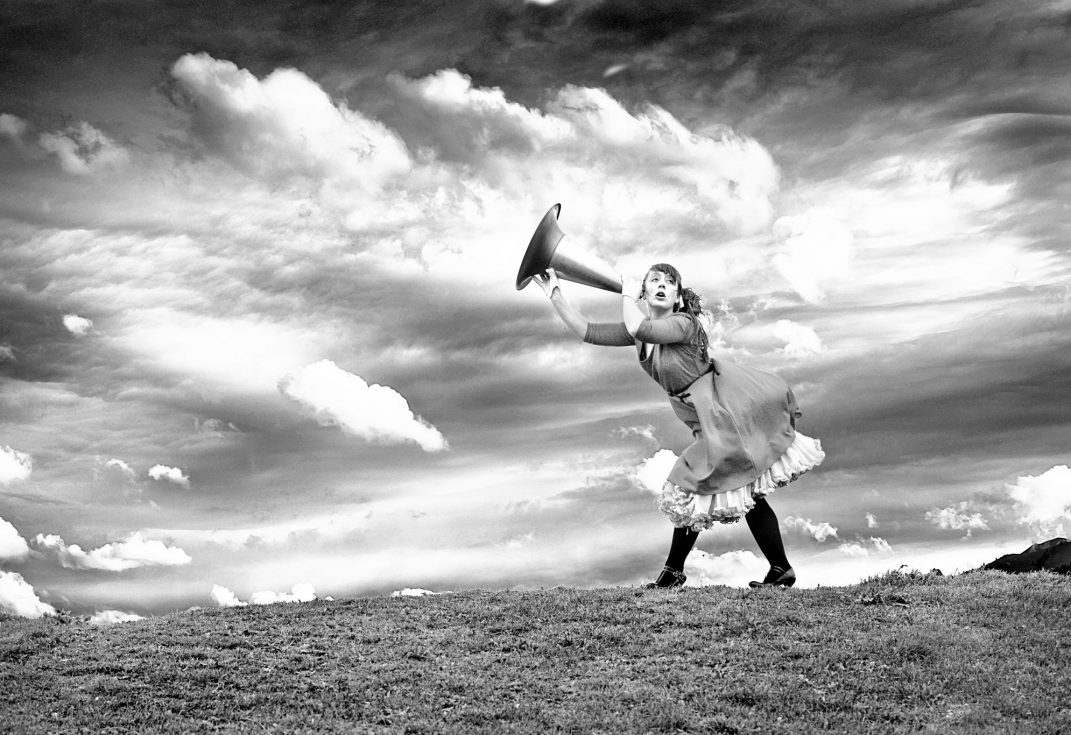 Black and white photo of women in costume standing on top of hill holding a large ear trumpet, with dramatic clouds behind
