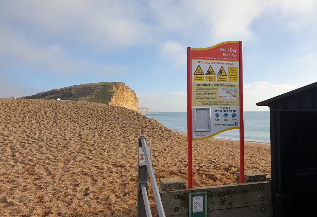 A warning sign in front of a shingle beach with cliffs and the sea in the background