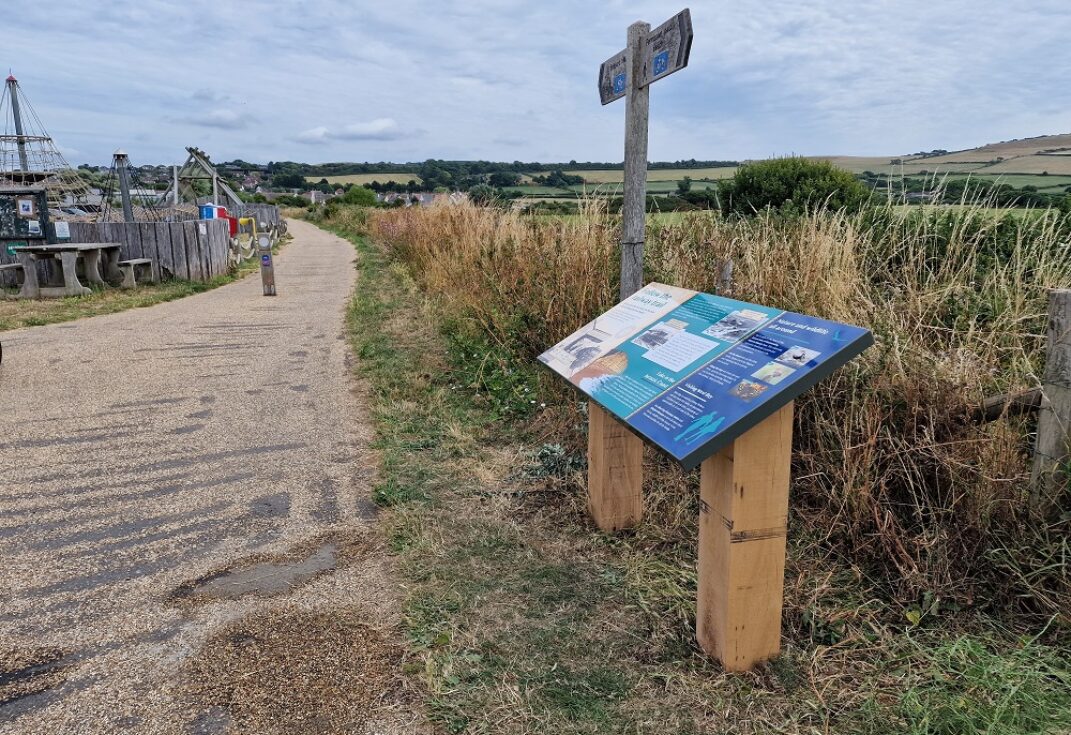An information board and signpost next to a gravel walkway next to a playground and surrounding fields