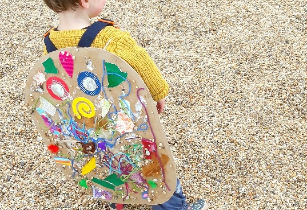 A child wearing a piece of art on their back standing on a shingle beach