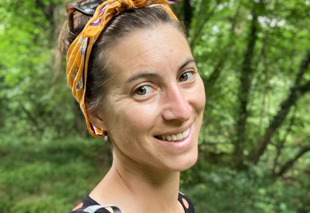 A woman with a headscarf and spotty top smiling at the camera with summer woodland behind