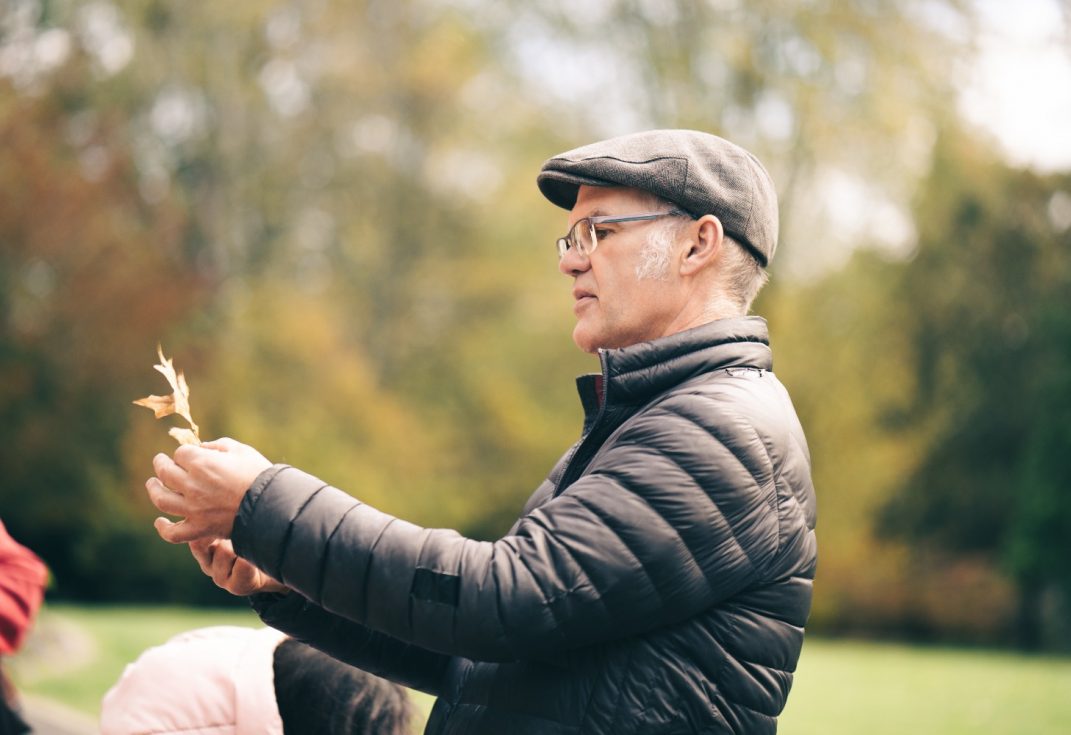 A man wearing glasses, a flat cap and winter jacket looking a a leaf outside with woods behind