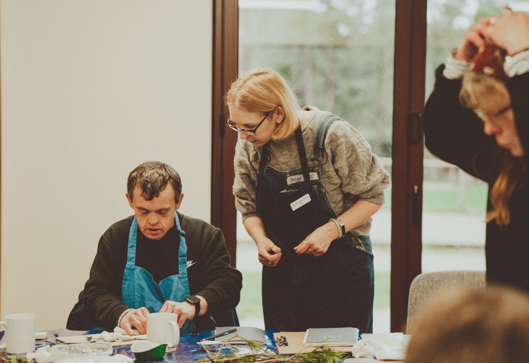 An art workshop inside with a man in an apron sitting working at a table and two women standing nearby watching and talking.