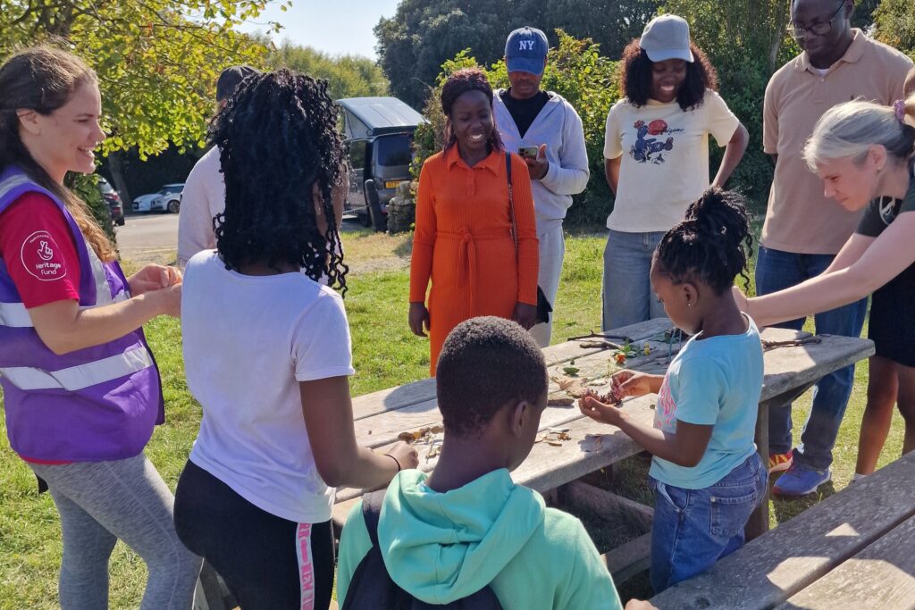A group of adults and children are gathered around an outdoor picnic table on a sunny day, taking part in a nature activity involving leaves and small natural objects. A facilitator wearing a purple vest stands with them, while others watch and participate under the shade of nearby trees.