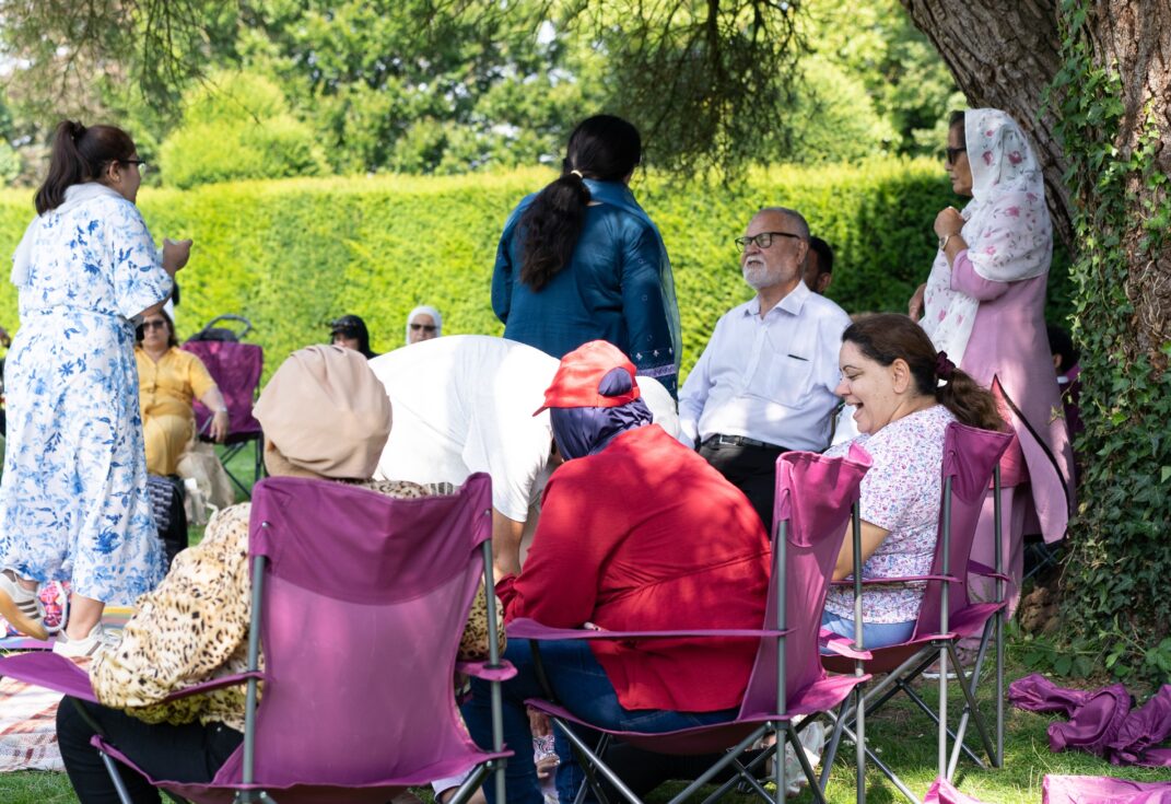 A group of adults are seated on camping chairs and picnic blankets in a grassy garden area, gathered under a large tree. Some people are chatting while others are standing or moving around. Pink and purple chairs are arranged in a semi-circle, and tall green hedges and trees form the background.