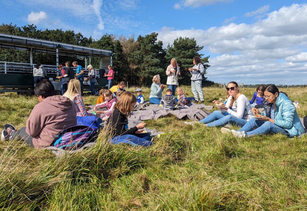 Picnic for Ukraine families on a visit to Middlebere Farm. Children and families sit on picnic blankets in a wide flat field, eating lunch, looking at the view of Poole Harbour. In the background is a farm trailer.