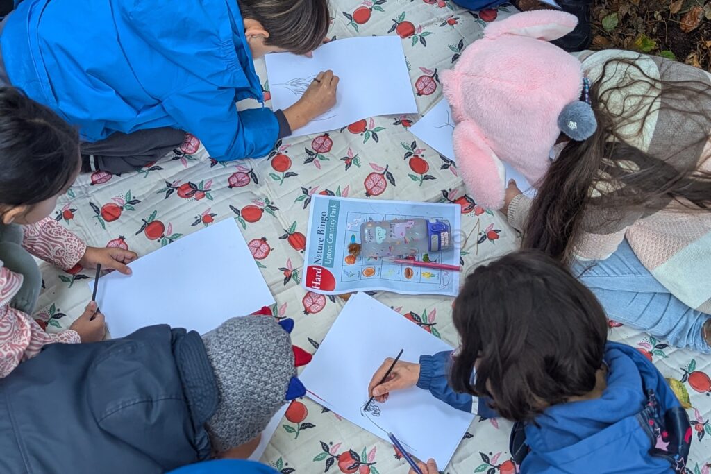 Children sitting on a patterned blanket outdoors, each drawing on sheets of white paper. A booklet titled “Field Sketching for Kids” lies in the centre, surrounded by pencils and sketchpads.