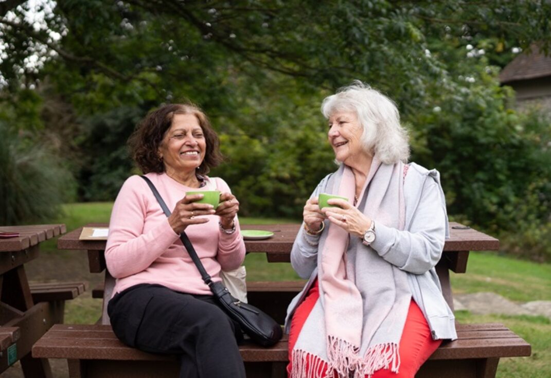 Two women sitting on a park bench laughing and having a cup of tea