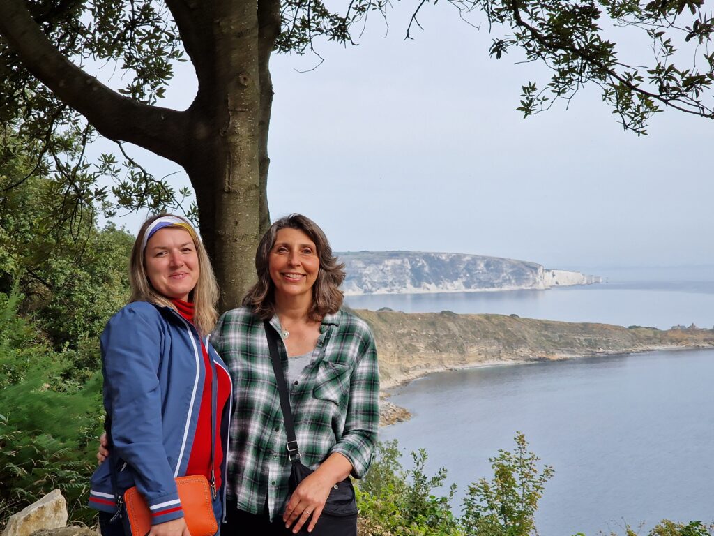 Two women pose in front of a tree with coastal cliffs and sea in the background