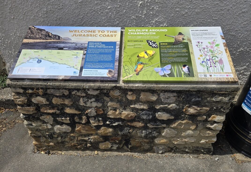 Two information boards on a stone pillar on a pavement next to a wall
