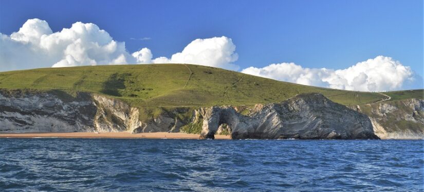 A view from the sea looking towards tall cliffs and an arch next to a beach on a sunny day