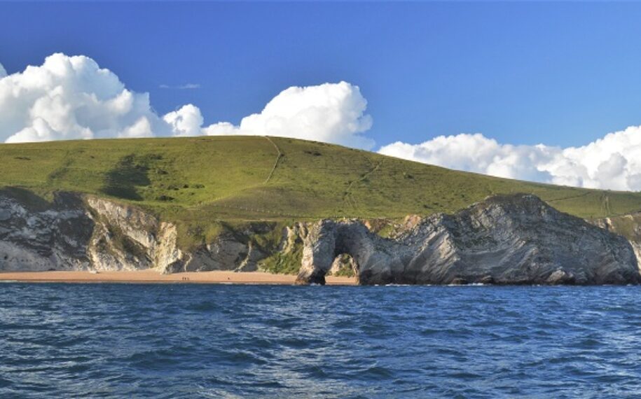 A view from the sea looking towards tall cliffs and an arch next to a beach on a sunny day