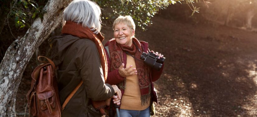 Two women enjoying time in a wood, one lady is holding binoculars and smiling.
