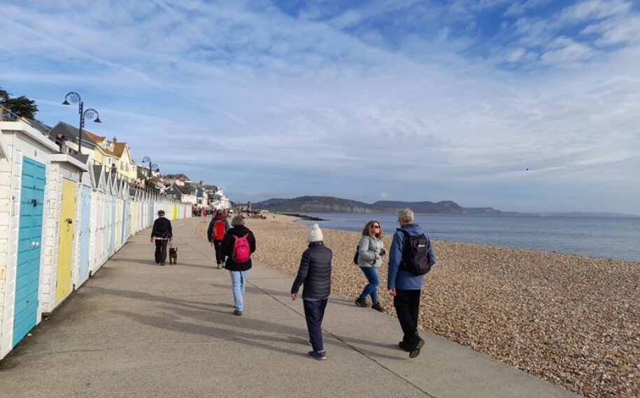 A group of walkers walk along the promenade , beach huts to left and shingle and sea to the right
