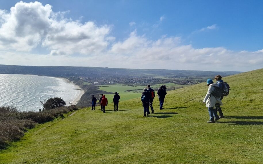 Group of walkers in outdoor clothes stop to admire the view across a wide bay from the coast path on a hill.