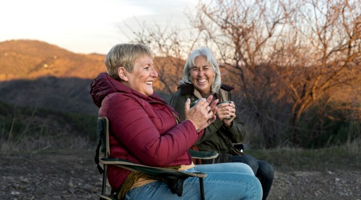 Two older women sit in chairs enjoying a coffee together in the outdoors at sunset.