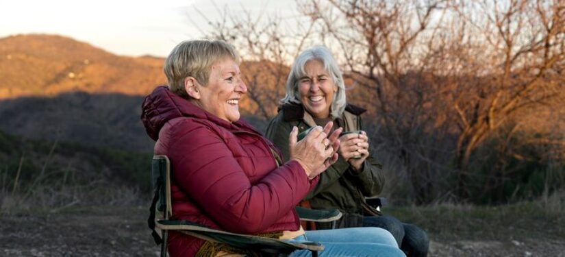 Two older women sit in chairs enjoying a coffee together in the outdoors at sunset.