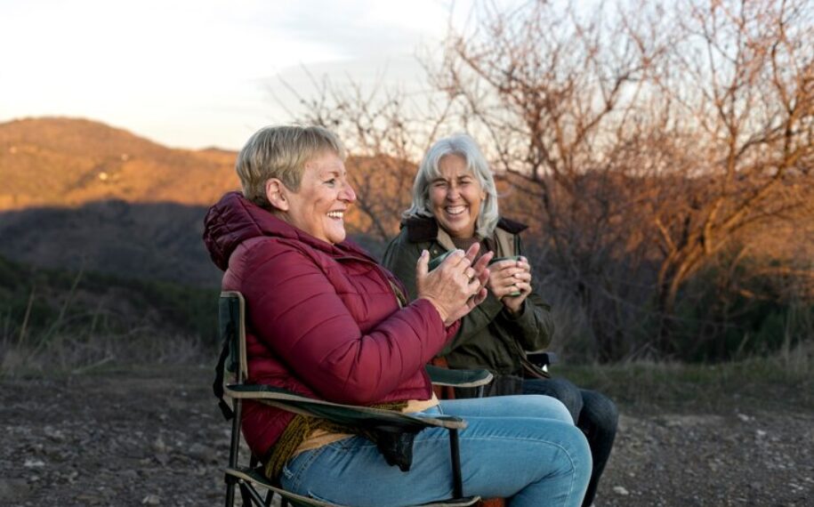 Two older women sit in chairs enjoying a coffee together in the outdoors at sunset.
