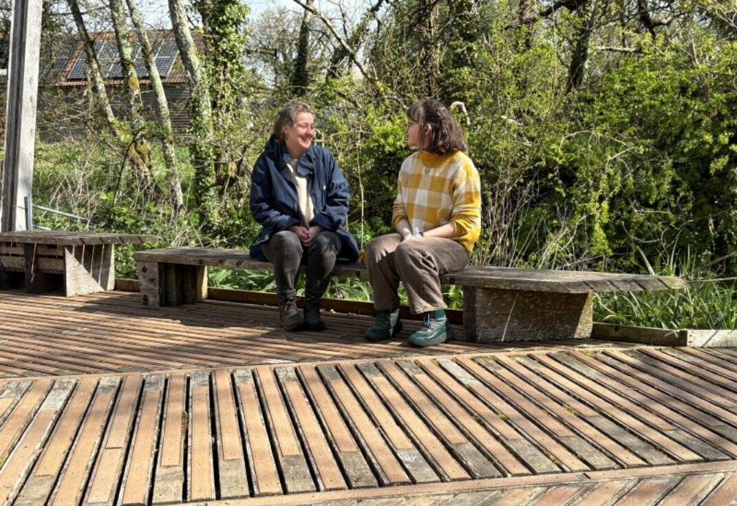 2 ladies sitting on wooden benching on a wooden board walk, backed by trees.