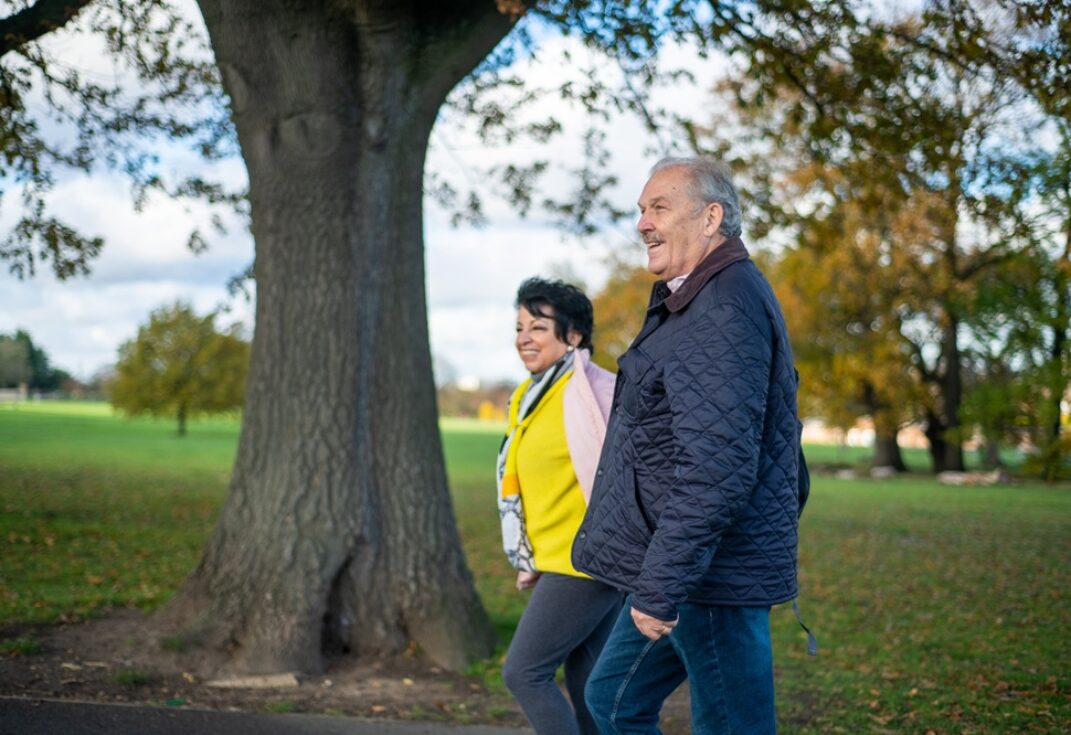 man and woman walking side by side in a park past a large tree.