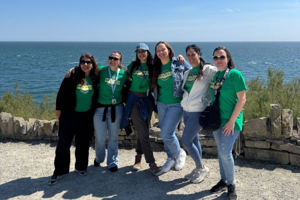 A group of six people stand side by side in front of a stone wall overlooking the sea on a sunny day. They are smiling and posing together, all wearing green T‑shirts, with the ocean and blue sky stretching out behind them.
