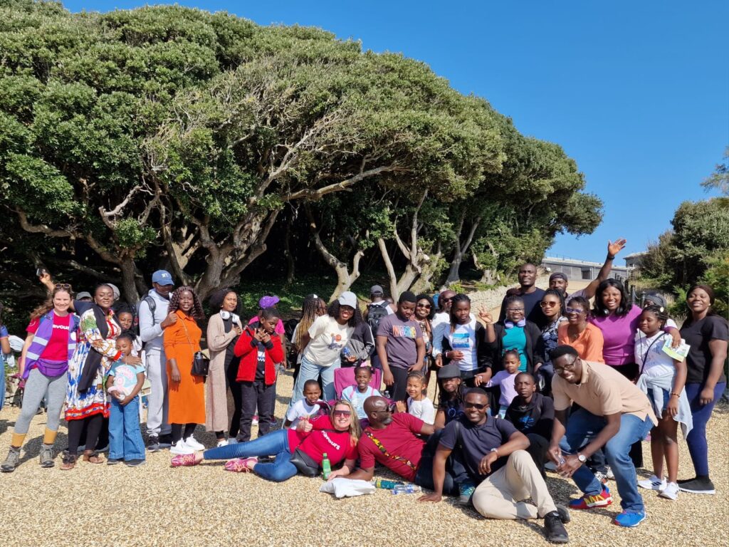 A large group of people of different ages gather outdoors on a bright sunny day. They stand and sit on a gravel area in front of a line of tall, windswept trees. Many are smiling and posing for a group photo.