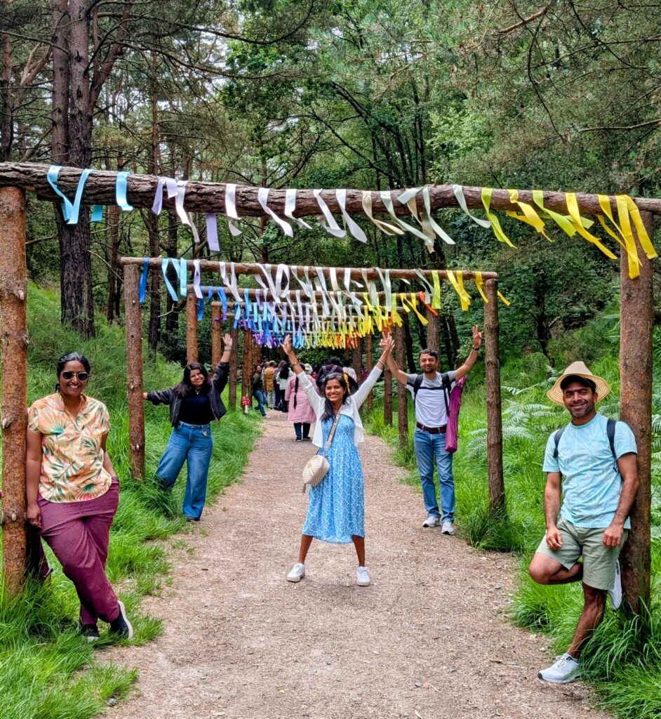 People pose playfully beneath a series of wooden archways decorated with colourful ribbons along a forest path. More people walk along the path behind them.