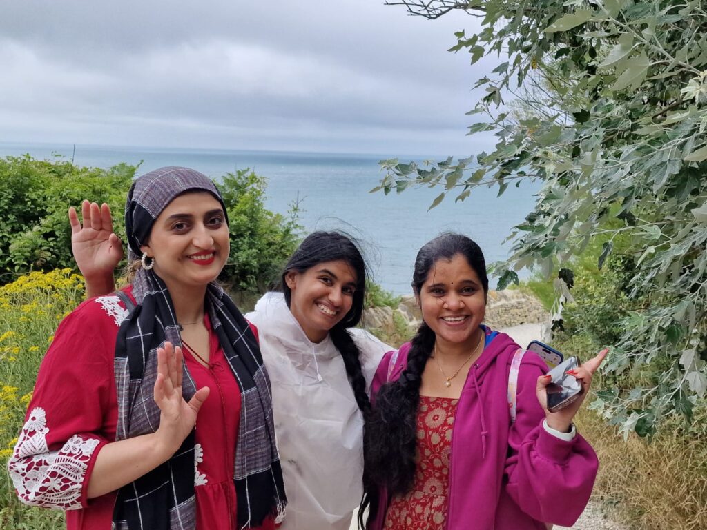 Three people stand together on a coastal path surrounded by greenery. The sea is visible behind them under a cloudy sky, and one person holds a phone.