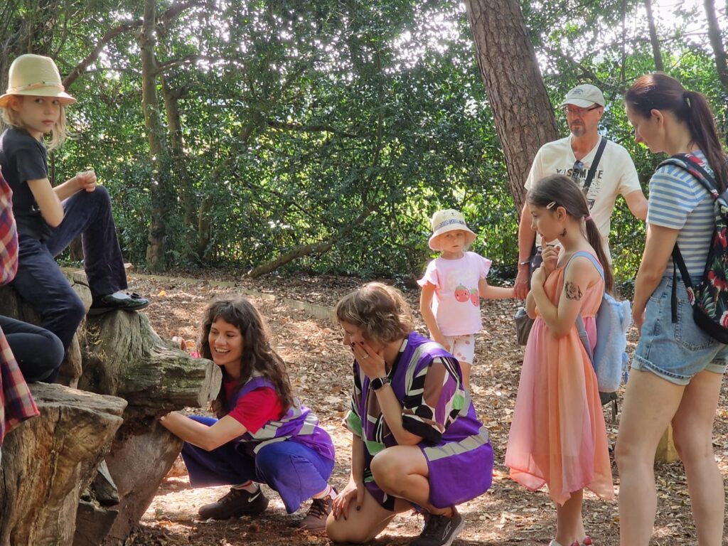 A group of children and adults gather in a wooded area. Some children crouch beside a fallen tree trunk while an adult and other children stand or sit nearby, all focused on something on the ground. Tall trees and dense greenery surround the group.