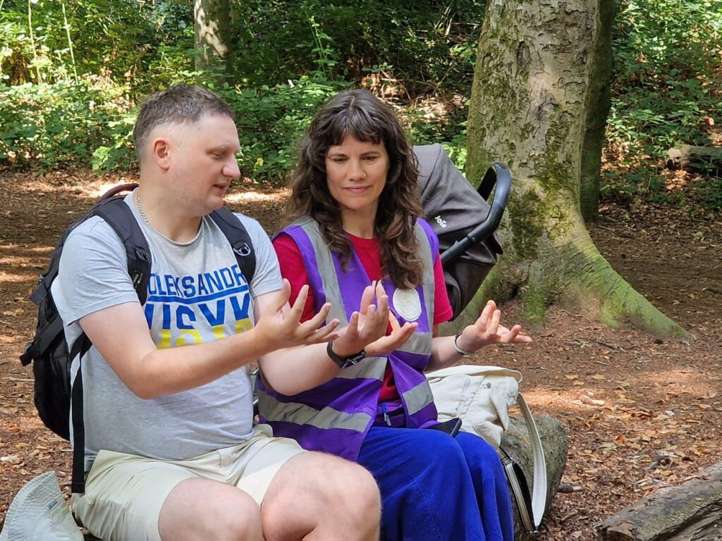 Two people sit on a log in a forested area, each holding their hands out in front of them in a mirrored gesture. One person wears a backpack, and a pushchair is visible behind them. Sunlight filters through the trees.