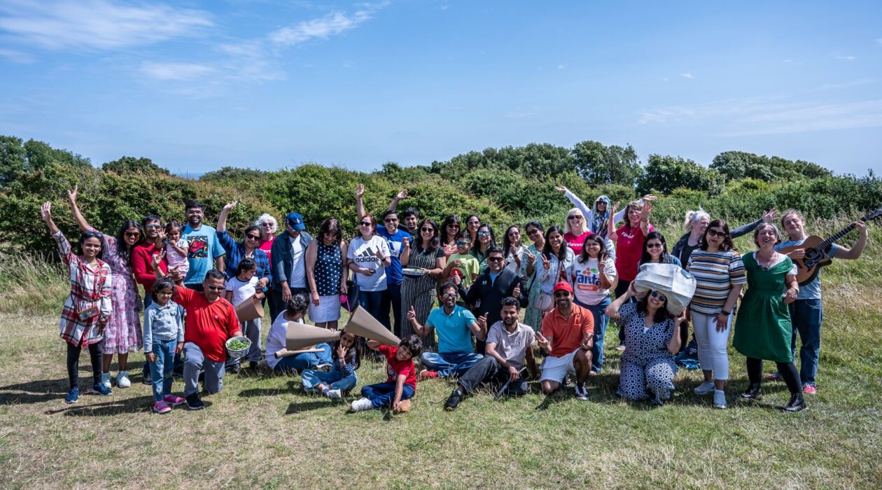 A large group of adults and children pose together in an open grassy area on a sunny day. Some hold props. Bushes and blue sky form the backdrop.