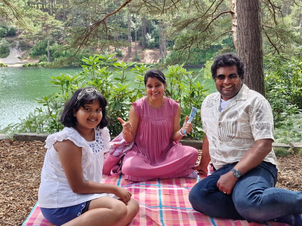A family sits on a blanket in a shaded forest area beside a lake. Trees and green shrubs surround them, with the water visible behind.
