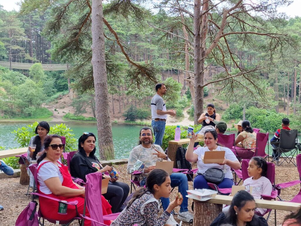 People sit in camping chairs and on the ground enjoying food at a lakeside picnic area shaded by tall pine trees. A lake and wooded hillside are visible behind them.