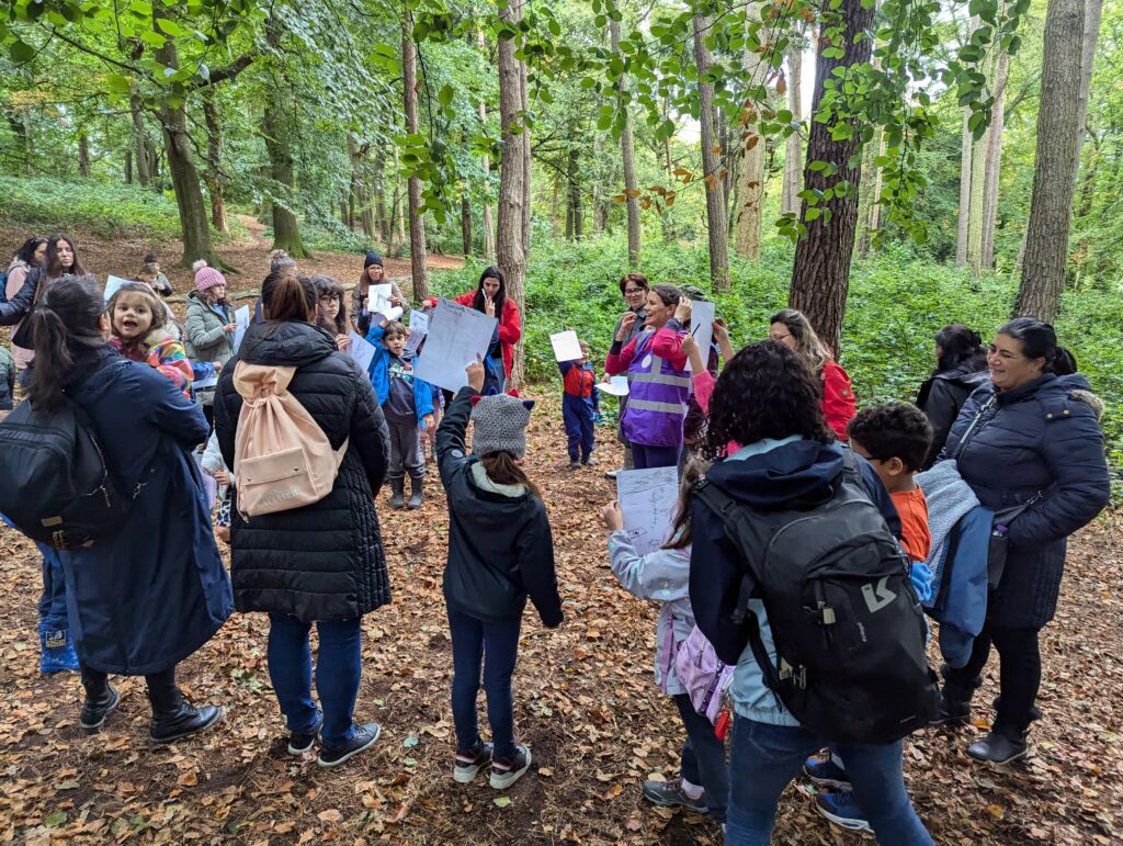 A group of adults and children stand in a wooded area surrounded by tall trees and green foliage, taking part in an outdoor learning activity. Several participants hold up sheets of paper or drawings while gathered in a circle on a leaf-covered forest floor. The scene is set along a woodland path, with dense vegetation and natural light filtering through the trees.