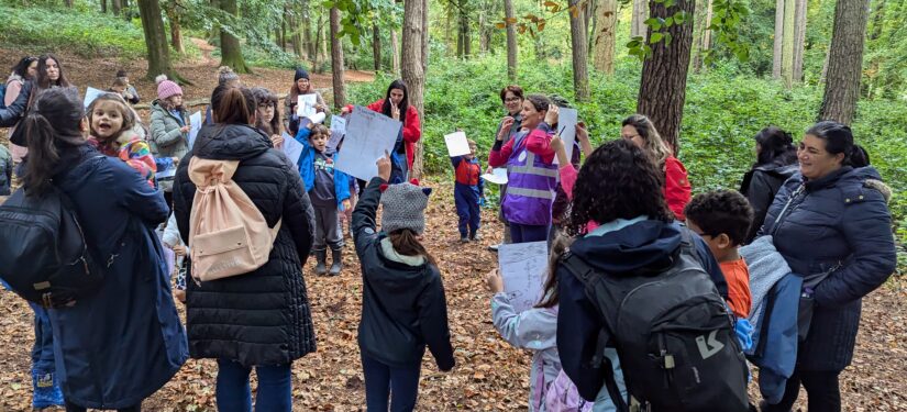A group of adults and children stand in a wooded area surrounded by tall trees and green foliage, taking part in an outdoor learning activity. Several participants hold up sheets of paper or drawings while gathered in a circle on a leaf-covered forest floor. The scene is set along a woodland path, with dense vegetation and natural light filtering through the trees.
