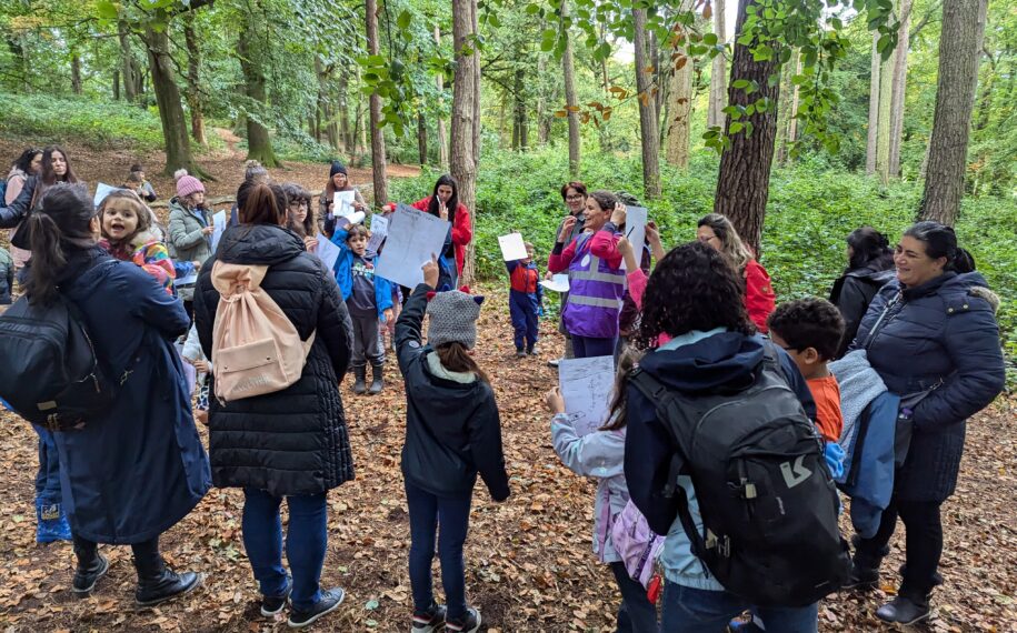 A group of adults and children stand in a wooded area surrounded by tall trees and green foliage, taking part in an outdoor learning activity. Several participants hold up sheets of paper or drawings while gathered in a circle on a leaf-covered forest floor. The scene is set along a woodland path, with dense vegetation and natural light filtering through the trees.