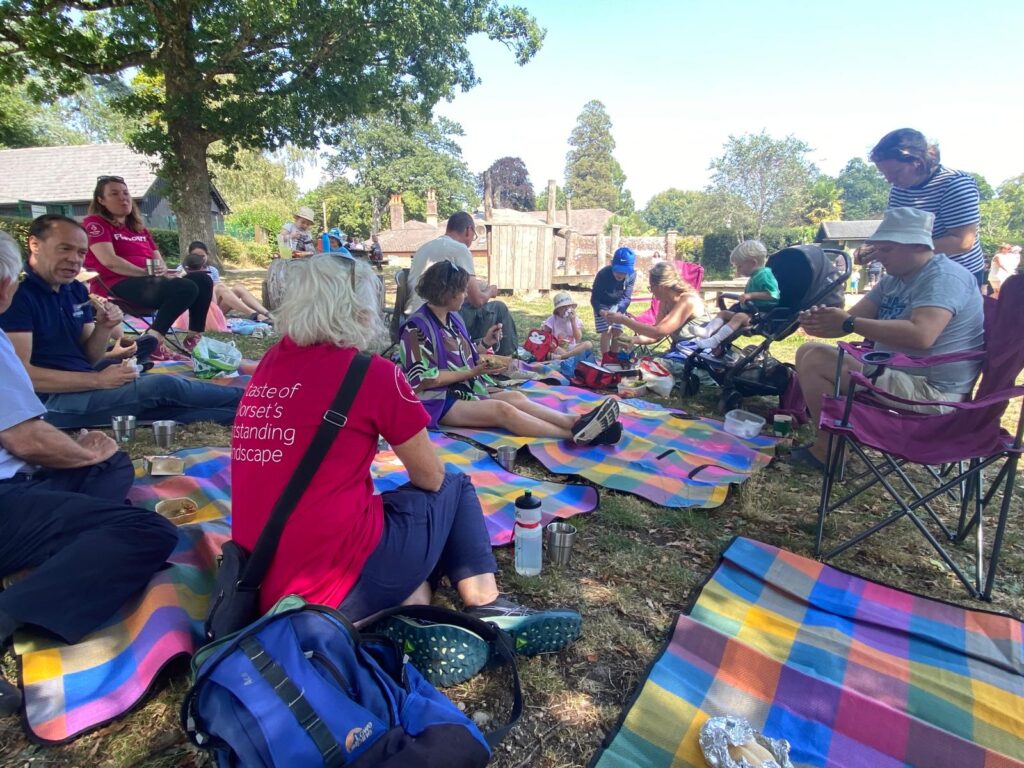 People sit together on colourful striped picnic blankets under the shade of large trees. Some sit on camping chairs while others relax on the blankets, chatting and eating. Bags, water bottles, and food containers are scattered around. A few buildings and trees are visible in the background.