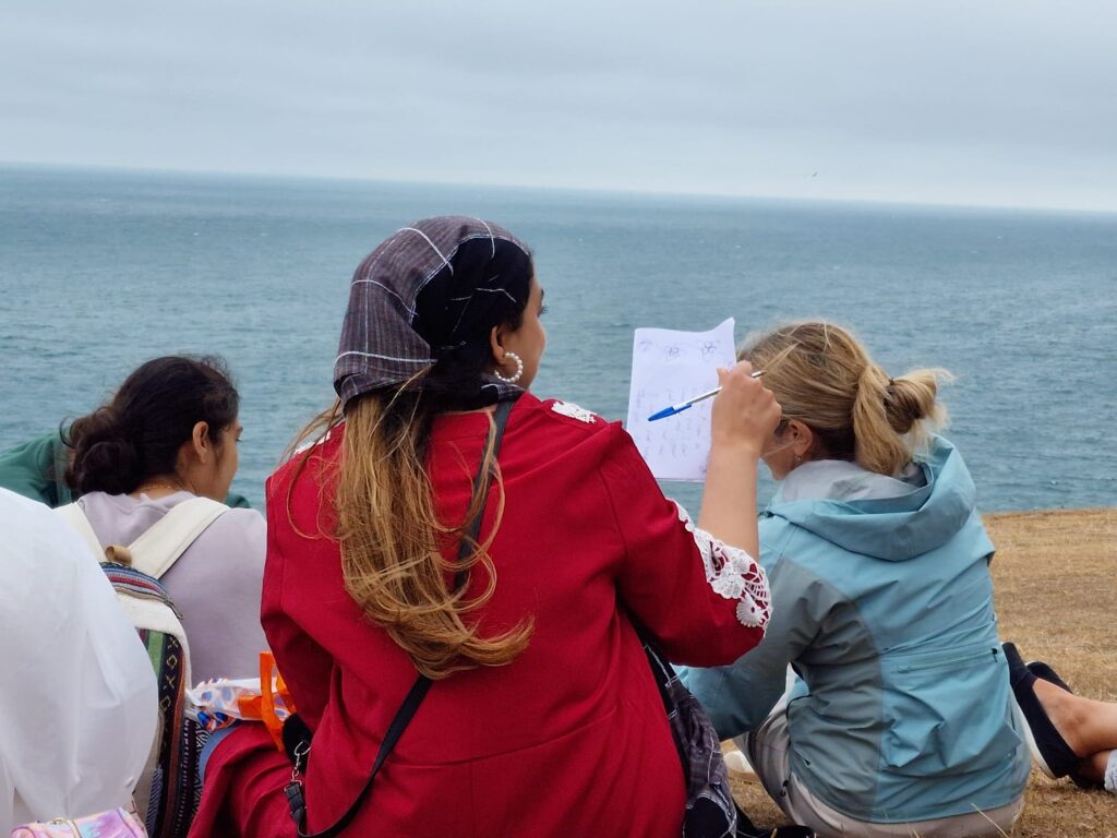 Three people sit on a grassy coastal cliff overlooking the sea. One person in a red top holds up a piece of paper with drawings while others look toward the water under an overcast sky.