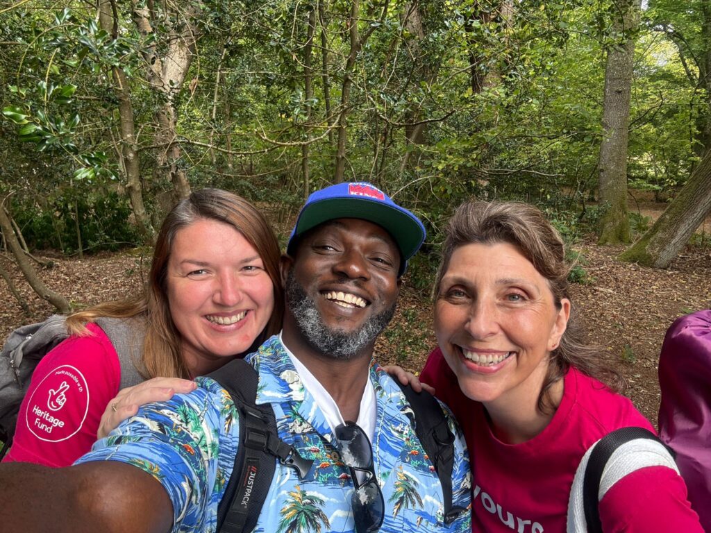 Three people gather closely for a selfie in a woodland area. They wear outdoor clothing and backpacks, with trees and forest floor visible in the background.