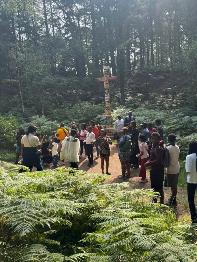 A large group of people stand on a forest path gathered around a tall, colourful totem pole. The area is surrounded by dense ferns and tall trees, with sunlight filtering through the canopy.
