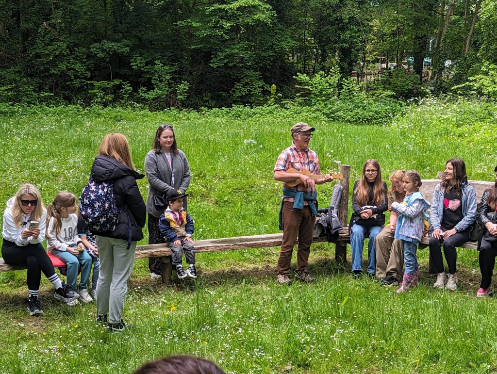 Children and adults sit on wooden benches in a grassy woodland clearing. An adult stands in front of the group holding a ukulele, while others listen or watch.