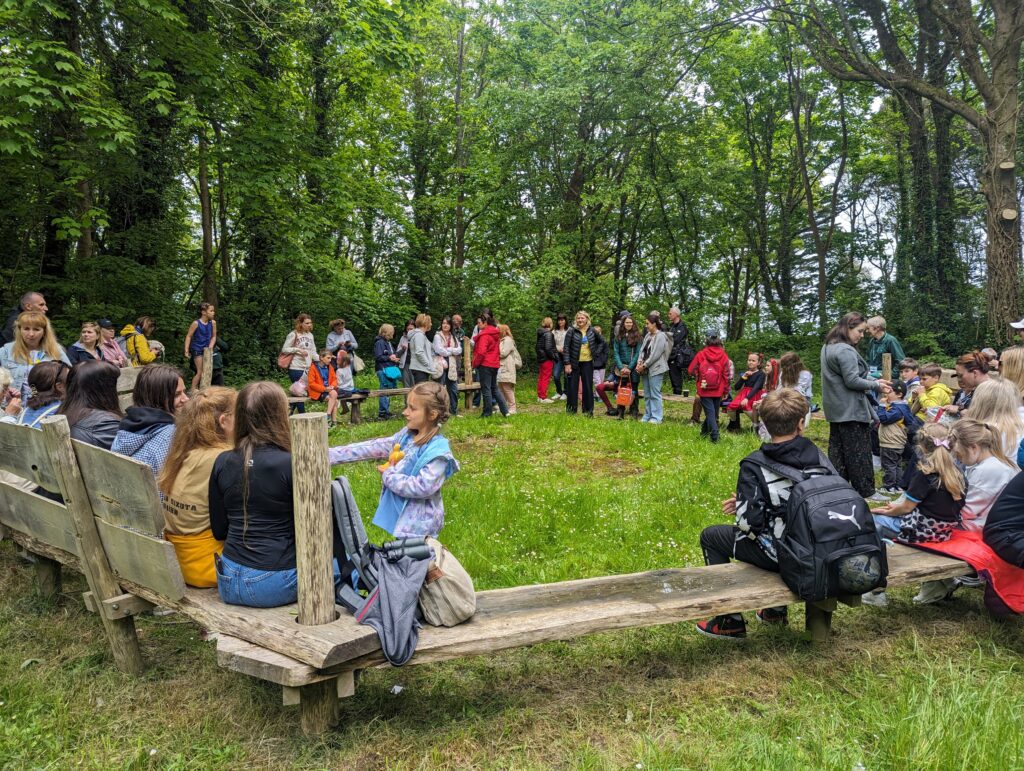A large group of children and adults gather in a clearing in a woodland area. Many sit on wooden benches arranged in a circle, while others stand and talk among the trees.