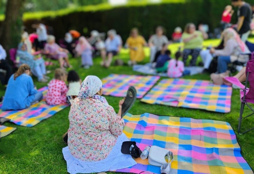 A group of people sit on colourful checked picnic blankets laid out on a grassy area in a park. Some individuals are seated on the blankets while others sit on folding chairs. The scene appears to be a relaxed outdoor gathering under the shade of a large tree, with bags, water bottles, and personal items placed around the blankets.
