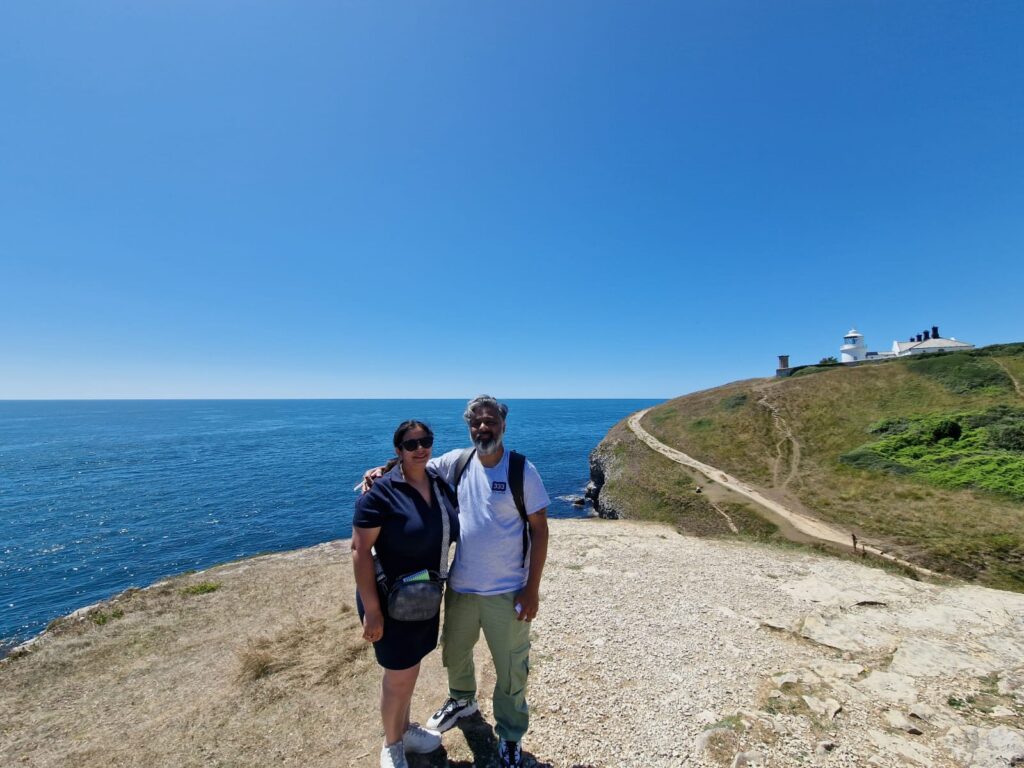 Two people standing close together on a coastal cliff path with the sea behind them. The background shows deep blue water and a rocky coastline under a clear sky.