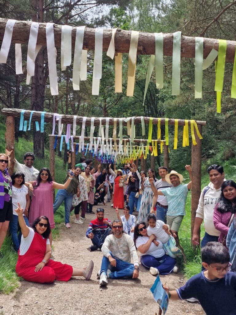 A large group of people sitting and standing along a decorated woodland walkway, with colourful hanging ribbons attached to wooden beams overhead.
