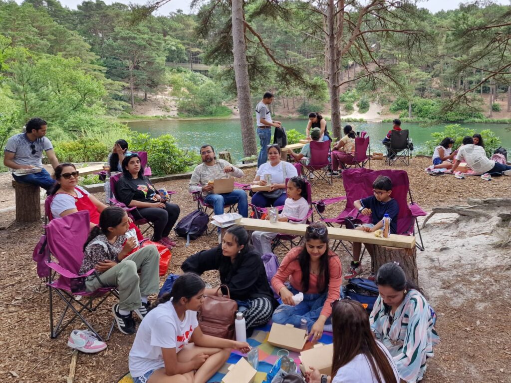 A group of people sitting in camping chairs having a picnic beside a lake, with tall trees and water in the background