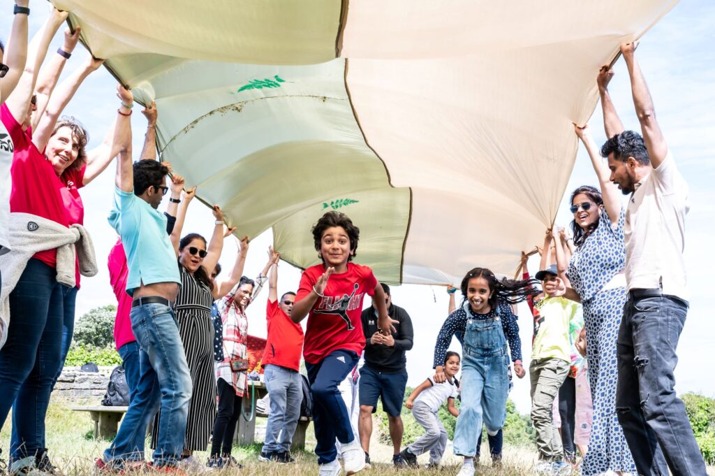 A group of adults forming an arch overhead with a large fabric sheet while several children run underneath it on a grassy field.