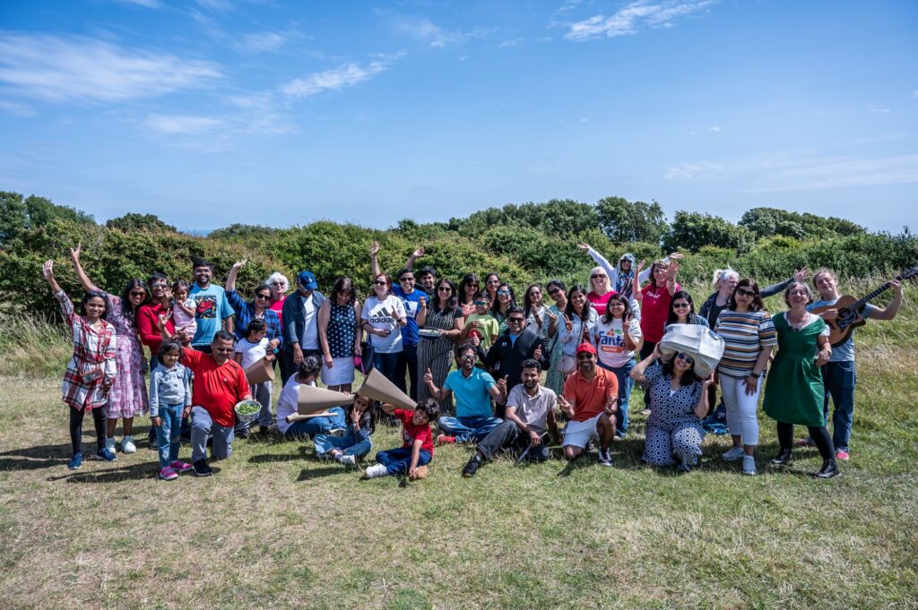 A large group of people outdoors on a grassy field, posing for a group photo. Some people are standing while others are sitting on the ground, with trees and blue sky in the background.
