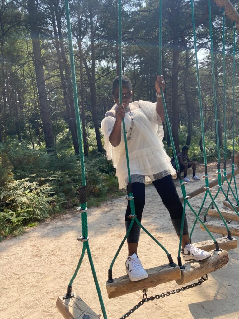 A person stands on a wooden swing-style bridge in a forest activity area, holding the ropes on either side.